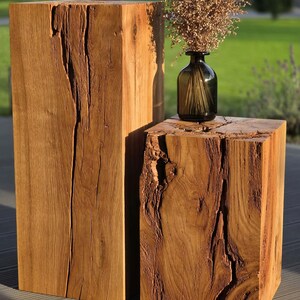 Op de afbeelding: Twee rechthoekige houten blokken met natuurlijke nerven, diepe scheuren en een ruwe textuur. Op het kortere houten blok staat een kleine, donkerglazen fles met gedroogde, lichtbruine botanische stengels.
