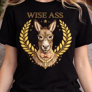 May include: Black t-shirt featuring a detailed illustration of a donkey wearing glasses, a bow tie, and a pearl necklace, surrounded by a golden laurel wreath. The text "WISE ASS" is arched above the donkey.