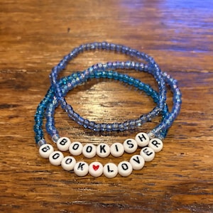 May include: Three blue beaded bracelets. One bracelet spells out "BOOKISH LOVER" in white letter beads with a red heart. The other two bracelets are made of blue beads.