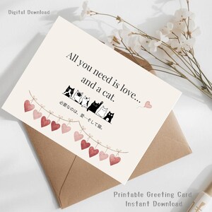 以下が含まれることがあります： 「All you need is love... and a cat.」のテキストが入ったグリーティングカード。水彩画のハートの列と、イラストの猫の列が特徴です。茶色の封筒に入っています。