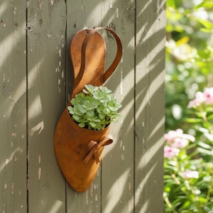 Puede incluir: Una jardinera de metal marrón oxidado con forma de zapato, con una correa y un lazo, llena de plantas verdes. Está montada en una valla de madera verde desgastada, creando un encantador acento de jardín.