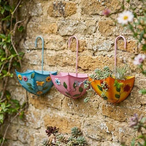 May include: Three decorative, umbrella-shaped wall planters in blue, pink, and orange. Each planter is adorned with floral and butterfly motifs. The planters are filled with plants and mounted on a textured stone wall, adding a touch of colour.