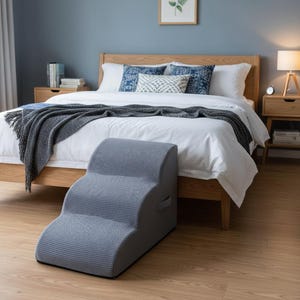 May include: A grey pet step with three steps, positioned in front of a bed. The step has a textured, patterned surface and a handle. The bed features a white duvet, blue cushions, and a wooden frame. The room has wooden flooring.