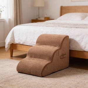 Puede incluir: Un escalón marrón para mascotas con tres niveles, diseñado para ayudar a perros o gatos pequeños a alcanzar superficies más altas. Tiene una cubierta de tela texturizada y un asa. Se encuentra junto a una cama.