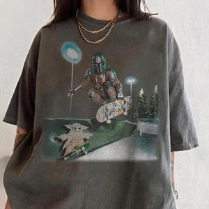 May include: Dark gray t-shirt featuring a graphic of The Mandalorian and Grogu skateboarding in a skate park. The design includes a night scene with streetlights and trees. The shirt has a vintage, worn look.