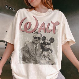 May include: Cream-colored t-shirt featuring a vintage-style graphic of Walt Disney and Mickey Mouse. The word "Walt" is printed in a pink, cursive font above the image. The image is a black and white photograph of Walt Disney and Mickey Mouse.