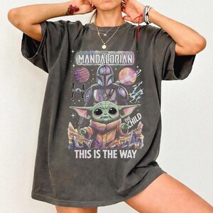 Pode incluir: Camiseta cinza escuro com um gr&aacute;fico de The Mandalorian. O design inclui o Mandaloriano, The Child, planetas e o texto "THIS IS THE WAY". A camiseta tem mangas curtas e um corte folgado.