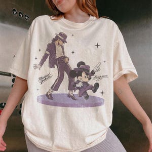 May include: Cream-colored t-shirt featuring a graphic of Michael Jackson and Mickey Mouse in matching purple outfits. The text "Michael Jackson" and "Mickey Mouse" are also visible. The shirt has a relaxed fit and short sleeves.