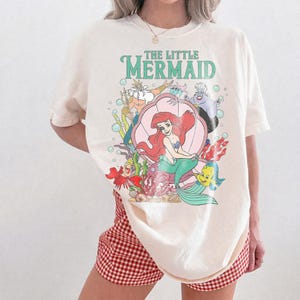 May include: Cream-colored t-shirt featuring a colorful graphic of Ariel from The Little Mermaid. The graphic includes the text "THE LITTLE MERMAID" in green, along with other characters from the movie. The shirt is paired with red and white checkered shorts.