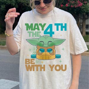K&ouml;nnte beinhalten: Cremefarbenes T-Shirt mit einer Grafik eines gr&uuml;nen Alien-Charakters, der zwei blaue Tassen h&auml;lt. Der Text "MAY THE 4TH BE WITH YOU" ist oberhalb und unterhalb der Figur in Gr&uuml;n, Blau und Braun gedruckt.