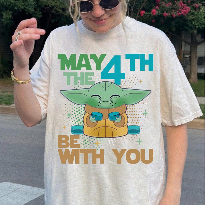 K&ouml;nnte beinhalten: Cremefarbenes T-Shirt mit einer Grafik eines gr&uuml;nen Alien-Charakters, der zwei blaue Tassen h&auml;lt. Der Text "MAY THE 4TH BE WITH YOU" ist oberhalb und unterhalb der Figur in Gr&uuml;n, Blau und Braun gedruckt.