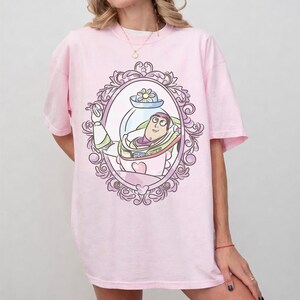 May include: A light pink t-shirt featuring a framed illustration of Buzz Lightyear from Toy Story. The illustration is in a decorative oval frame with a heart and flower. The shirt has short sleeves and a crew neck.
