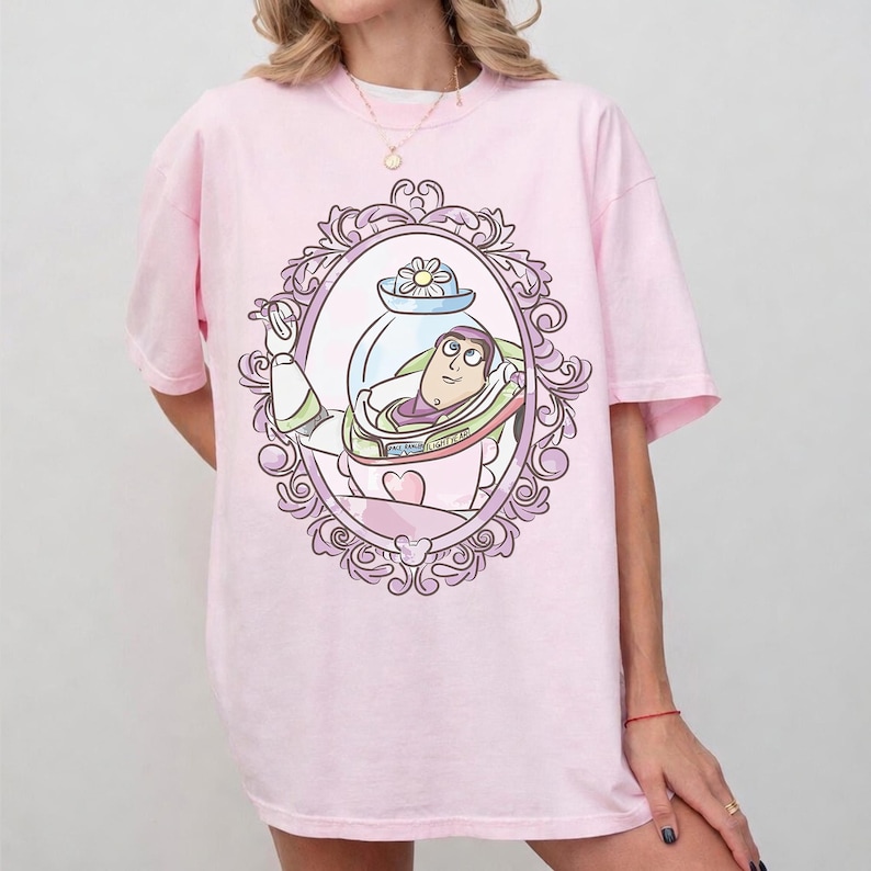 May include: A light pink t-shirt featuring a framed illustration of Buzz Lightyear from Toy Story. The illustration is in a decorative oval frame with a heart and flower. The shirt has short sleeves and a crew neck.