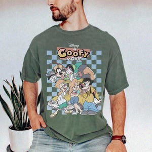 May include: Green t-shirt featuring a graphic from Disney's "A Goofy Movie." The design includes Goofy and other characters in a colorful, cartoon style. The shirt has a checkered background and the movie title in a retro font.