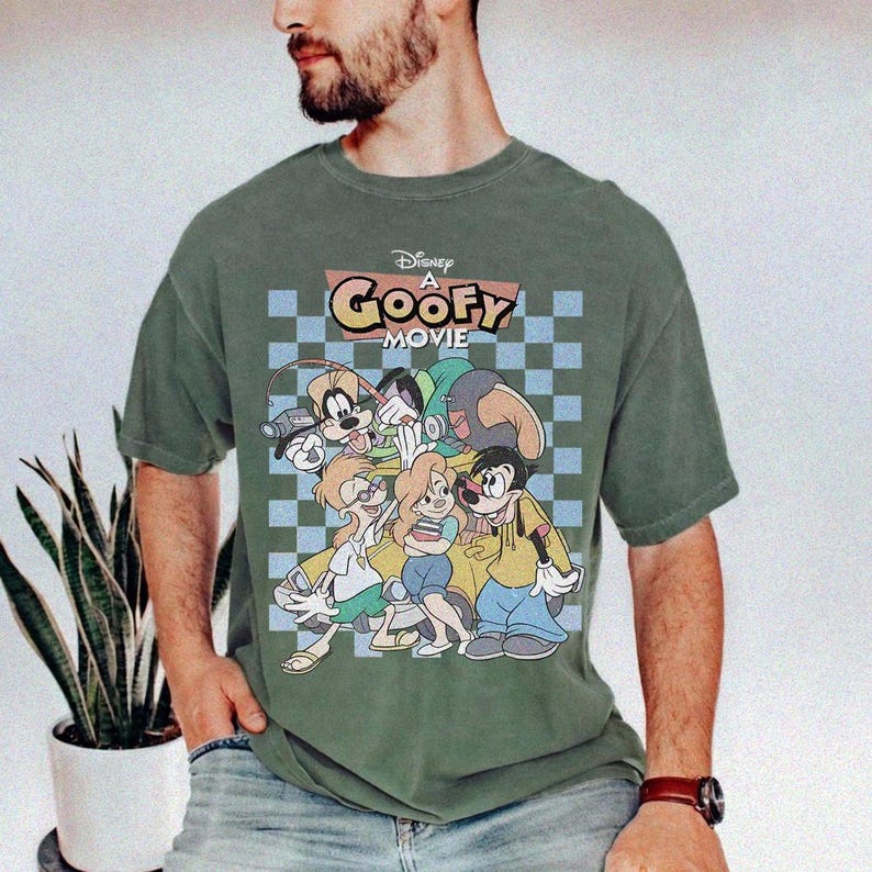 May include: Green t-shirt featuring a graphic from Disney's "A Goofy Movie." The design includes Goofy and other characters in a colorful, cartoon style. The shirt has a checkered background and the movie title in a retro font.
