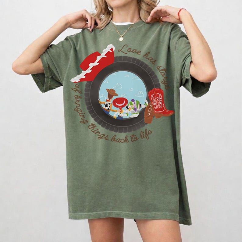 May include: An olive green t-shirt featuring a graphic of a tire framing cartoon characters. The design includes a red cowboy hat, a red boot with the name Taylor, and the text Love has stories of bringing things back to life in brown.
