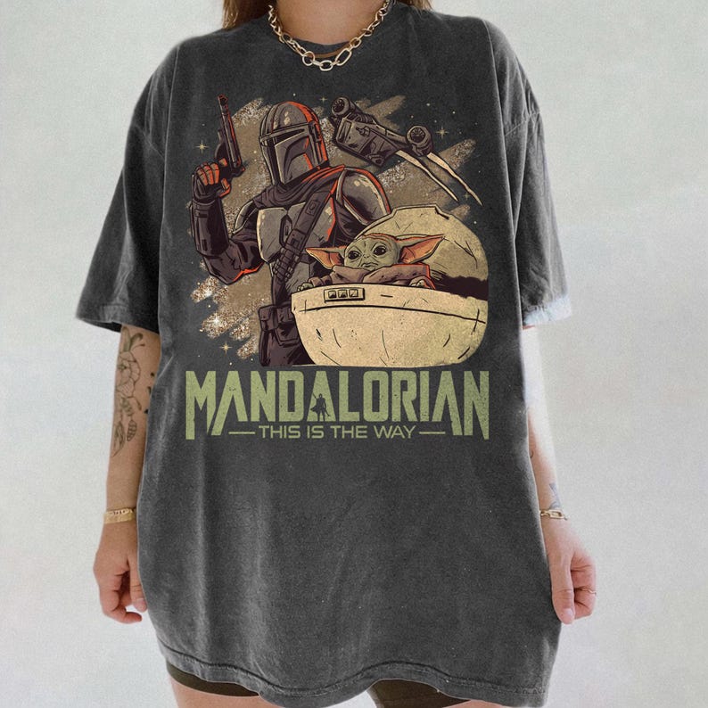 May include: A charcoal gray oversized t-shirt featuring a graphic print of a Mandalorian character holding a blaster and a small creature in a floating pod. The text "MANDALORIAN THIS IS THE WAY" is printed in large green letters below the illustration.