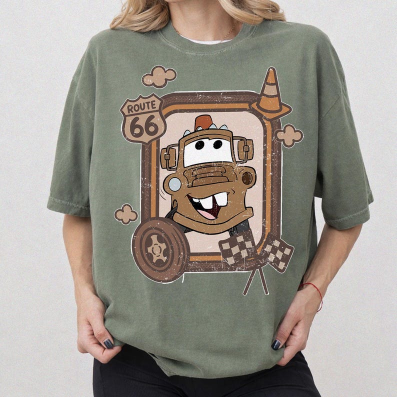 May include: Olive green t-shirt featuring a cartoon image of Tow Mater from the movie Cars. The design includes a Route 66 sign, a traffic cone, a tire, and a checkered flag. The graphic is in shades of brown, beige, and white.