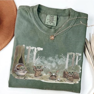 May include: Green Comfort Colors t-shirt featuring a Grogu graphic in a forest scene. The design includes Grogu in a pod, playing with a ball, and drinking from a cup. A gold necklace rests on the shirt. The shirt has a crew neck and short sleeves.