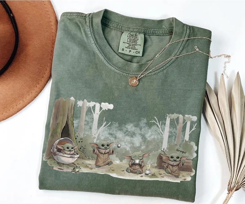 May include: Green Comfort Colors t-shirt featuring a Grogu graphic in a forest scene. The design includes Grogu in a pod, playing with a ball, and drinking from a cup. A gold necklace rests on the shirt. The shirt has a crew neck and short sleeves.
