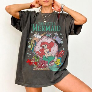 May include: Dark gray t-shirt featuring a colorful graphic from "The Little Mermaid." The design includes Ariel, Flounder, Sebastian, and other characters. The words "The Little Mermaid" are printed above the graphic in green.