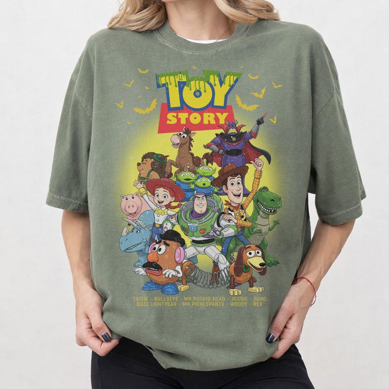 May include: An olive green crew neck t-shirt featuring a colourful graphic print of Toy Story characters including Woody, Buzz Lightyear, Jessie, and others. The design includes the Toy Story logo, yellow bats, and character names listed at the bottom.