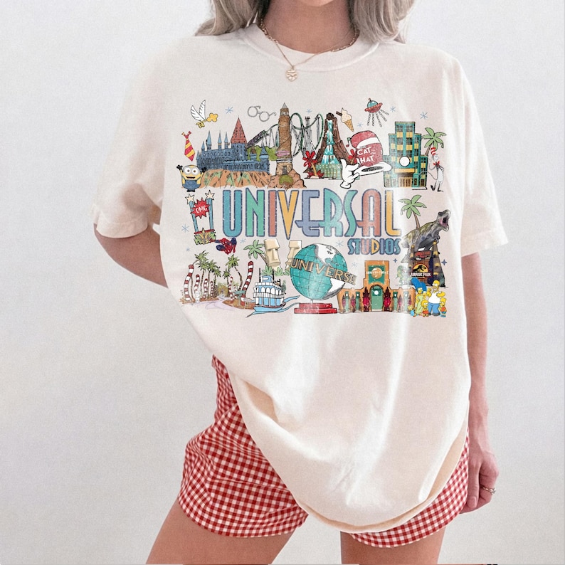 May include: Cream-colored t-shirt featuring a colorful graphic of Universal Studios attractions. The design includes the text "UNIVERSAL STUDIOS" in a multi-colored font, along with various illustrations of theme park elements. The shirt is paired with red and white checkered shorts.