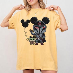 May include: Yellow t-shirt featuring a cartoon illustration of a Mandalorian character and Grogu, with a Mickey Mouse ear balloon. The Mandalorian is in gray armor with red accents, and Grogu is in a brown cloak. The design is on a yellow, short-sleeved shirt.