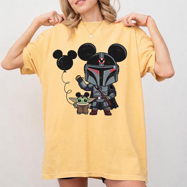 May include: Yellow t-shirt featuring a cartoon illustration of a Mandalorian character and Grogu, with a Mickey Mouse ear balloon. The Mandalorian is in gray armor with red accents, and Grogu is in a brown cloak. The design is on a yellow, short-sleeved shirt.