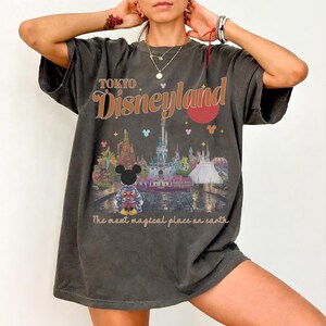 May include: Dark gray t-shirt featuring a graphic of Tokyo Disneyland with Mickey Mouse. The design includes the text "TOKYO Disneyland" in orange and the phrase "The most magical place on earth."