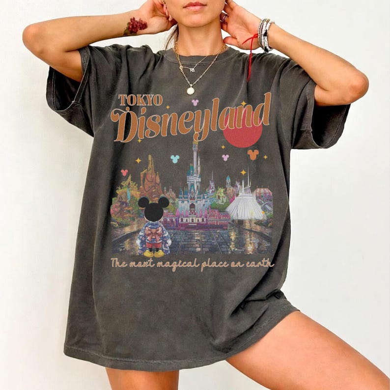 May include: Dark gray t-shirt featuring a graphic of Tokyo Disneyland with Mickey Mouse. The design includes the text "TOKYO Disneyland" in orange and the phrase "The most magical place on earth."