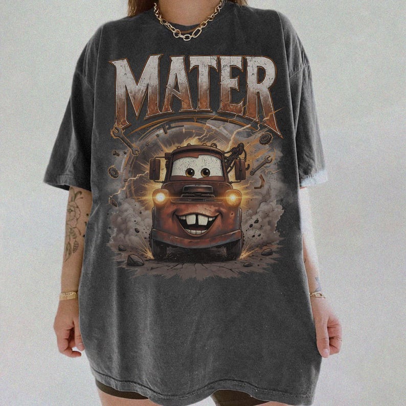 Puede incluir: Camiseta gris oscuro con una imagen de dibujos animados de Mater de la pel&iacute;cula Cars. La palabra "MATER" est&aacute; impresa en la parte superior en una fuente marr&oacute;n desgastada. El gr&aacute;fico incluye una ilustraci&oacute;n detallada de Mater con una cara sonriente, rodeada de elementos mec&aacute;nicos.