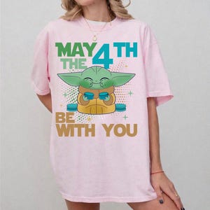 K&ouml;nnte beinhalten: Hellrosa T-Shirt mit einer Grafik eines gr&uuml;nen Alien-Charakters und dem Text "MAY THE 4TH BE WITH YOU". Das Design zeigt den Alien, der zwei blaue Objekte h&auml;lt. Der Text ist in Gr&uuml;n, Blau und Gold gehalten.