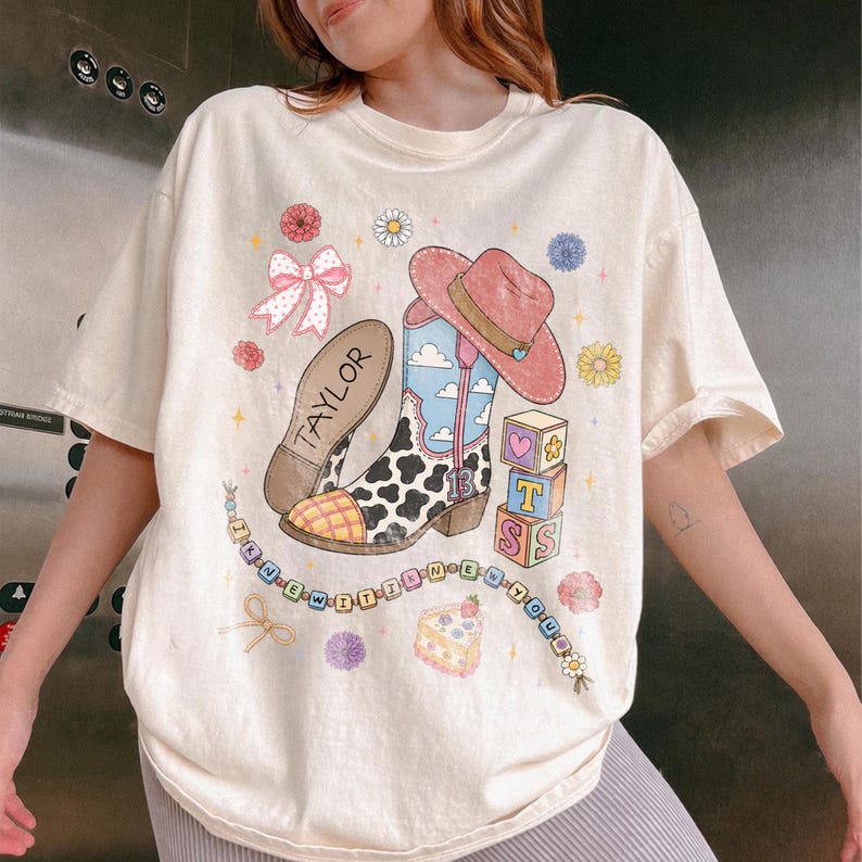 May include: A cream-colored t-shirt featuring a graphic print of a cow-print cowboy boot, a pink cowboy hat, a bow, alphabet blocks spelling T S, a slice of cake, and a beaded string with the text I KNEW IT WAS YOU and the name TAYLOR.