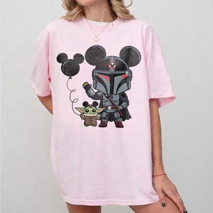 May include: A light pink t-shirt featuring a cartoon illustration of a Mandalorian character with a Baby Yoda figure. The Mandalorian is wearing a helmet with mouse ears. A black balloon with mouse ears is also in the image.