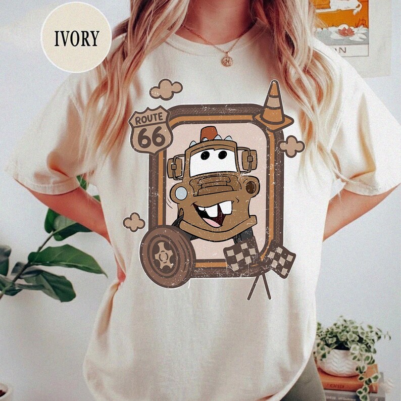 May include: Ivory-colored t-shirt featuring a cartoon image of Tow Mater from the movie Cars, framed by a Route 66 sign and a traffic cone. The design includes a wheel, clouds, and checkered flags. The word "IVORY" is printed on the top left.