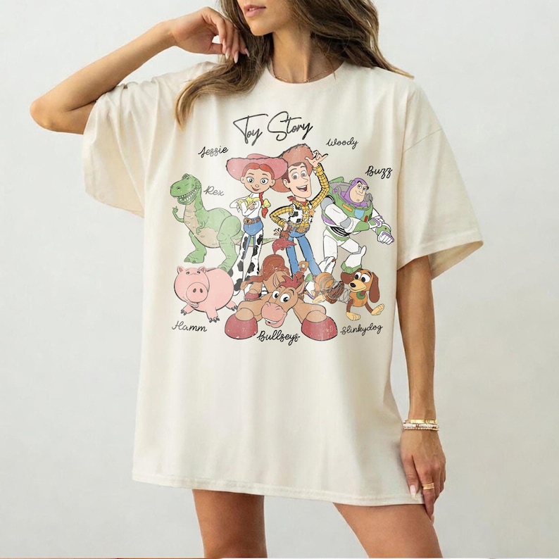 May include: Cream-colored t-shirt featuring the text "Toy Story" above a colorful graphic of characters including Woody, Buzz, Jessie, and Rex. The design includes Hamm, Bullseye, and Slinky Dog. The shirt has short sleeves.