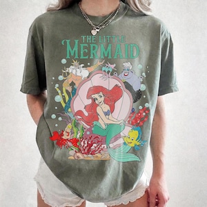 May include: Olive green t-shirt featuring a colorful graphic of Ariel from Disney's The Little Mermaid. The design includes Ariel in a seashell, King Triton, Flounder, and Ursula. The words "THE LITTLE MERMAID" are printed above the characters.