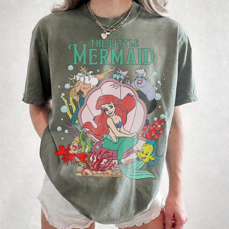 May include: Olive green t-shirt featuring a colorful graphic of Ariel from Disney's The Little Mermaid. The design includes Ariel in a seashell, King Triton, Flounder, and Ursula. The words "THE LITTLE MERMAID" are printed above the characters.