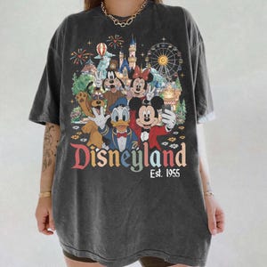 May include: A charcoal gray short-sleeve t-shirt featuring a graphic print of Mickey Mouse, Minnie Mouse, Donald Duck, Goofy, and Pluto taking a selfie in front of a castle and ferris wheel. The text reads Disneyland Est. 1955 in colorful letters.