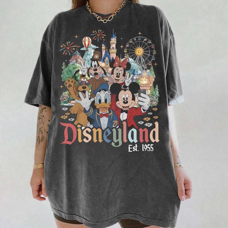 May include: A charcoal gray short-sleeve t-shirt featuring a graphic print of Mickey Mouse, Minnie Mouse, Donald Duck, Goofy, and Pluto taking a selfie in front of a castle and ferris wheel. The text reads Disneyland Est. 1955 in colorful letters.