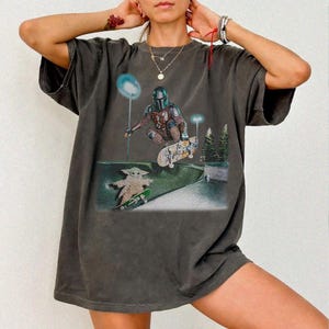 May include: Dark gray t-shirt featuring a graphic of The Mandalorian and Grogu skateboarding in a skate park. The design includes a detailed illustration of the characters and skateboards, set against a backdrop of a skate park with trees and lights.