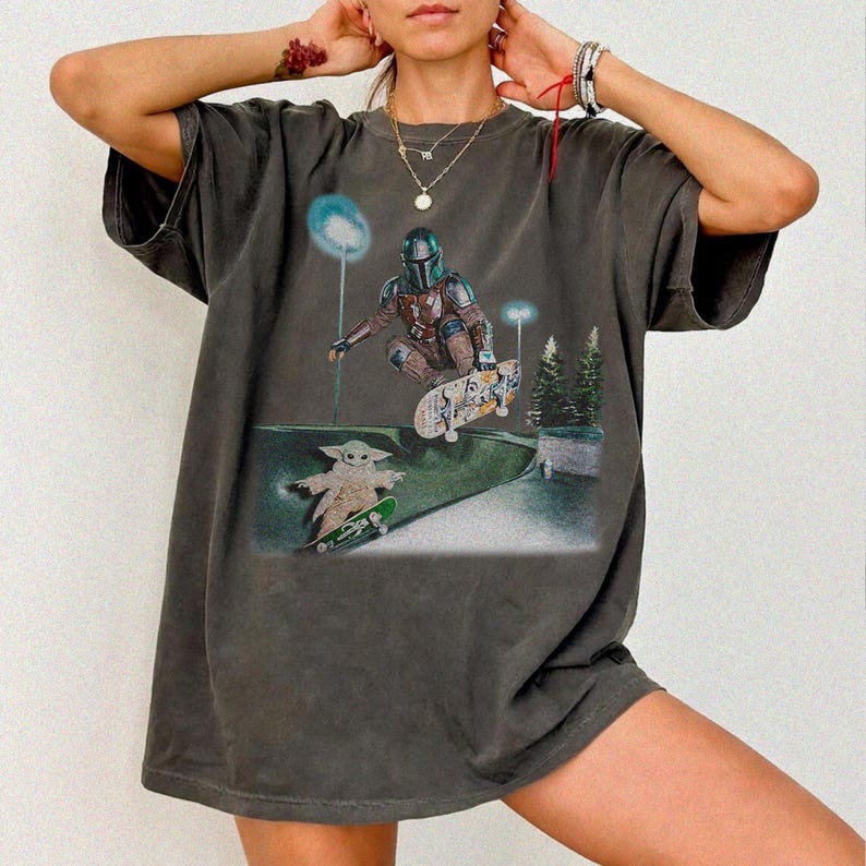 May include: Dark gray t-shirt featuring a graphic of The Mandalorian and Grogu skateboarding in a skate park. The design includes a detailed illustration of the characters and skateboards, set against a backdrop of a skate park with trees and lights.