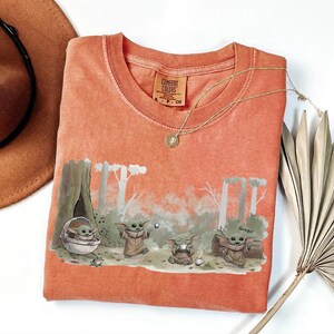 May include: Coral-colored t-shirt featuring a forest scene with Grogu characters. The graphic includes Grogu in a pod, playing with a ball, and sitting near a campfire. A gold necklace and a brown hat are also visible.
