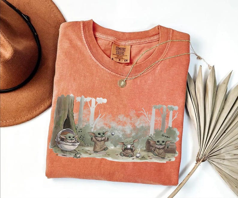 May include: Coral-colored t-shirt featuring a forest scene with Grogu characters. The graphic includes Grogu in a pod, playing with a ball, and sitting near a campfire. A gold necklace and a brown hat are also visible.