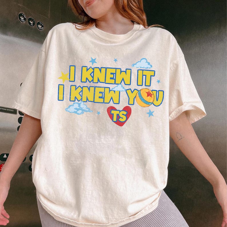 May include: A cream-colored short-sleeve t-shirt featuring the text "I KNEW IT I KNEW YOU" in bold yellow letters with blue outlines. The design includes a red heart with "TS" inside, a yellow ball with a red star, blue clouds, and small blue stars.