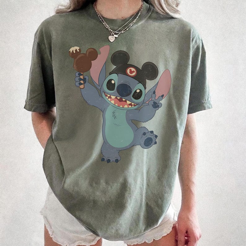 May include: Olive green t-shirt featuring a cartoon Stitch character wearing a Mickey Mouse hat and holding a Mickey Mouse-shaped ice cream bar. The character is blue with pink ears and a big smile. The shirt is a relaxed fit.