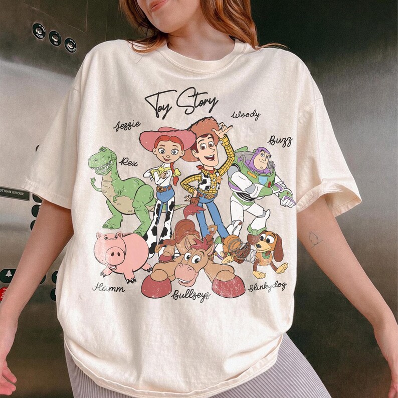 May include: Cream-colored t-shirt featuring the words "Toy Story" above a graphic of the movie's characters, including Woody, Buzz Lightyear, and Jessie. The shirt has a classic crew neck and short sleeves.