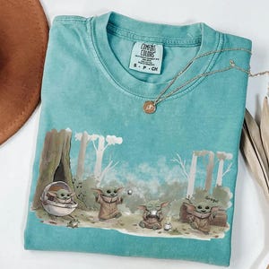 May include: A turquoise Comfort Colors t-shirt featuring a Grogu graphic. The design shows Grogu in various poses within a forest setting. A gold necklace with a pendant rests on the shirt. The shirt is size S-P-CH.