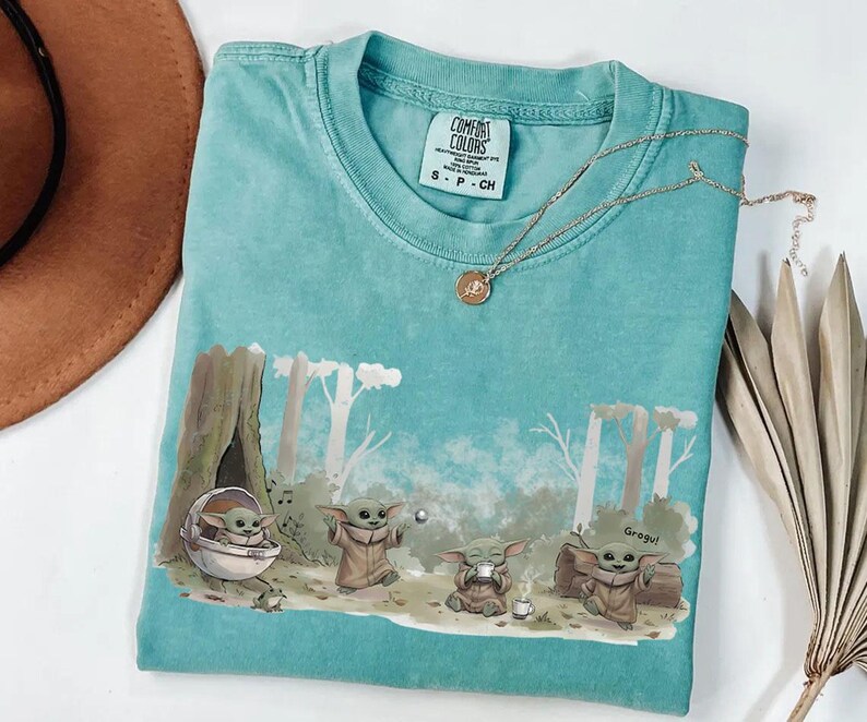 May include: A turquoise Comfort Colors t-shirt featuring a Grogu graphic. The design shows Grogu in various poses within a forest setting. A gold necklace with a pendant rests on the shirt. The shirt is size S-P-CH.